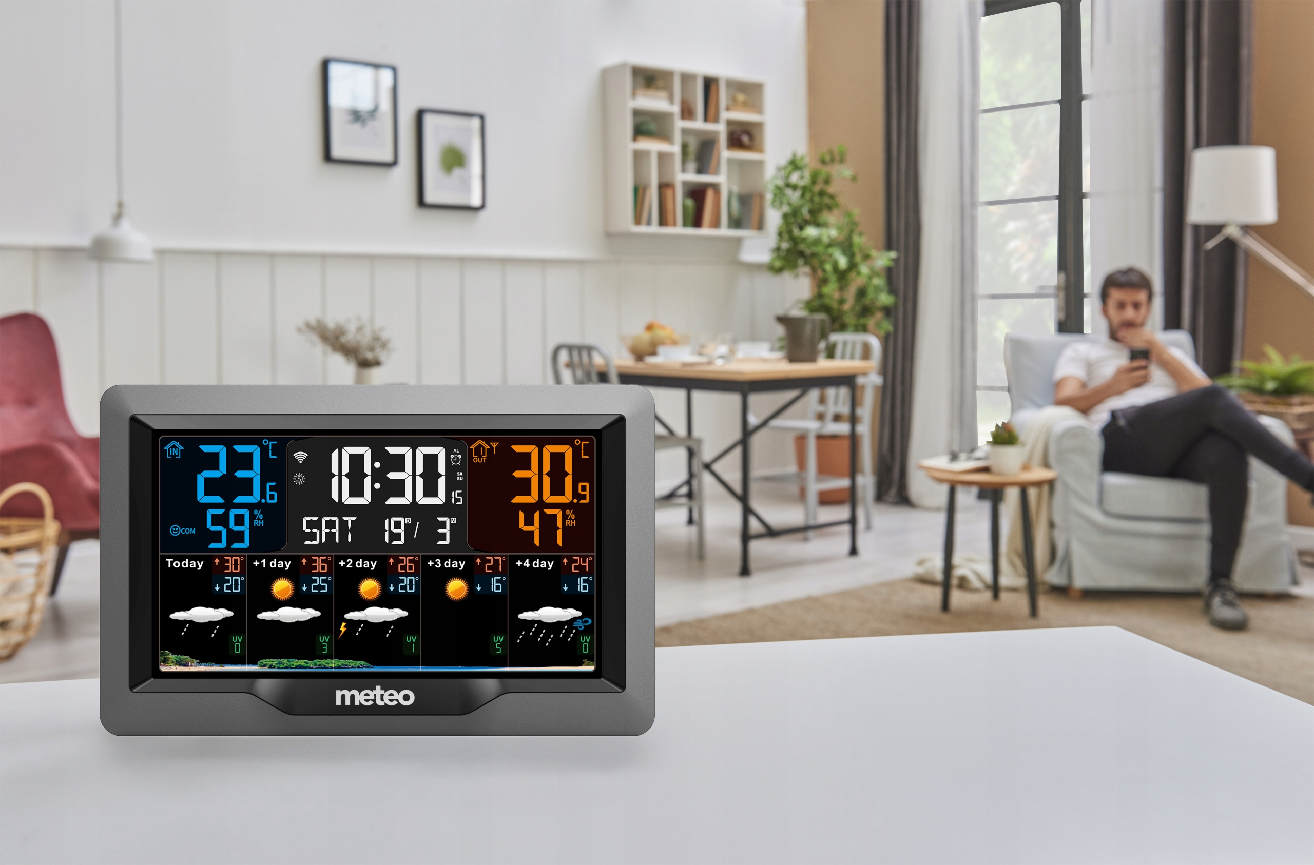 Stacja pogodowa wyświetla temperaturę i prognozę. Mężczyzna odpoczywa na kanapie w tle.