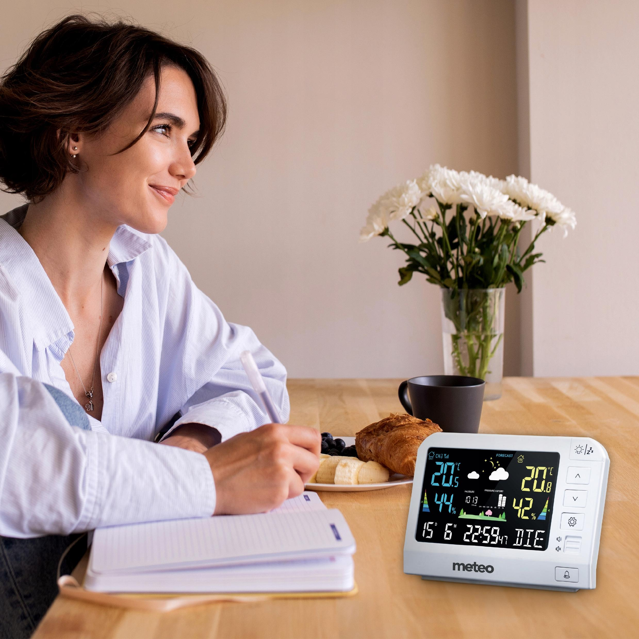 Kobieta uśmiecha się, pisze w notatniku, w pobliżu kwiatów, śniadania i stacji pogodowej. Stacja wyświetla temperaturę.