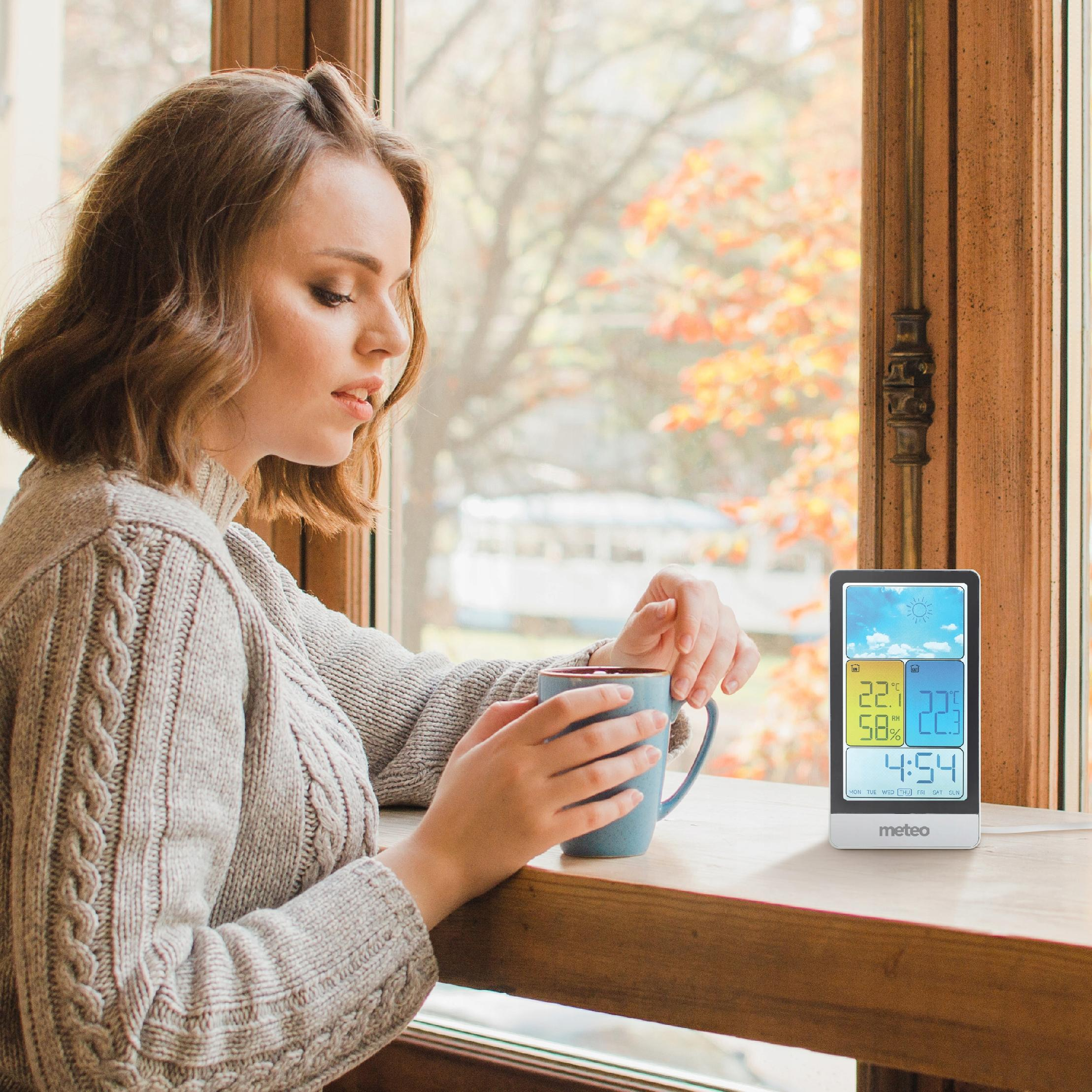 Kobieta trzyma niebieski kubek, siedząc przy oknie. Stacja pogodowa wyświetla czas i temperaturę. Widoczne są jesienne liście.
