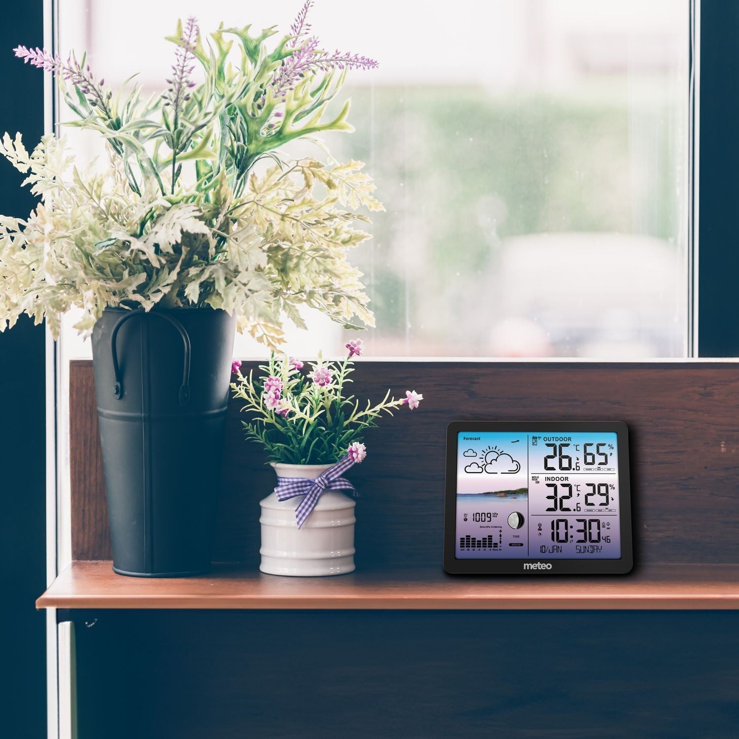 Półka ze stacją pogodową wyświetlającą temperaturę, godzinę i prognozę. Dwa wazony z kwiatami. W tle okno.