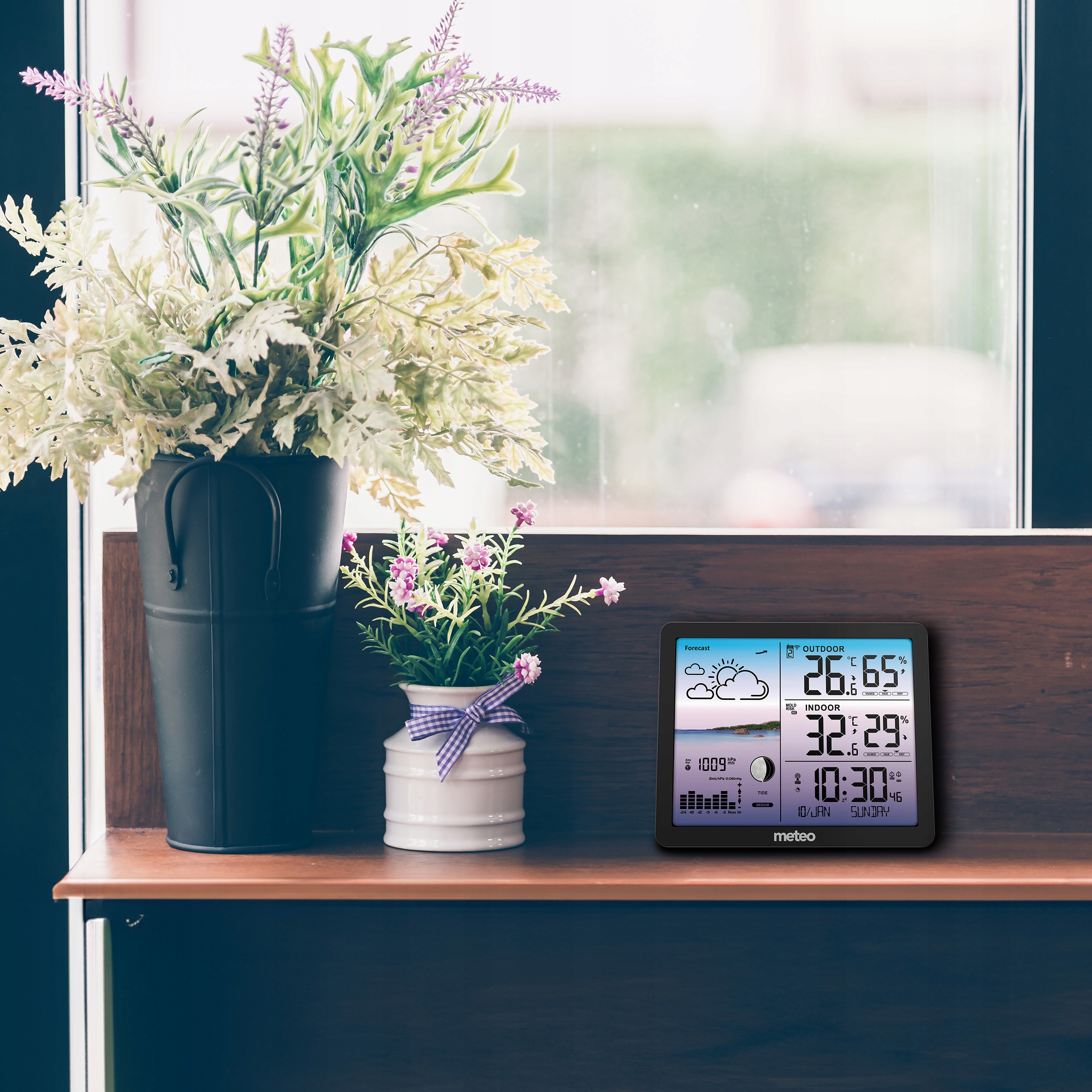 Stacja pogodowa wyświetla temperaturę i godzinę. Kwiaty w donicach stoją na półce. W tle okno.