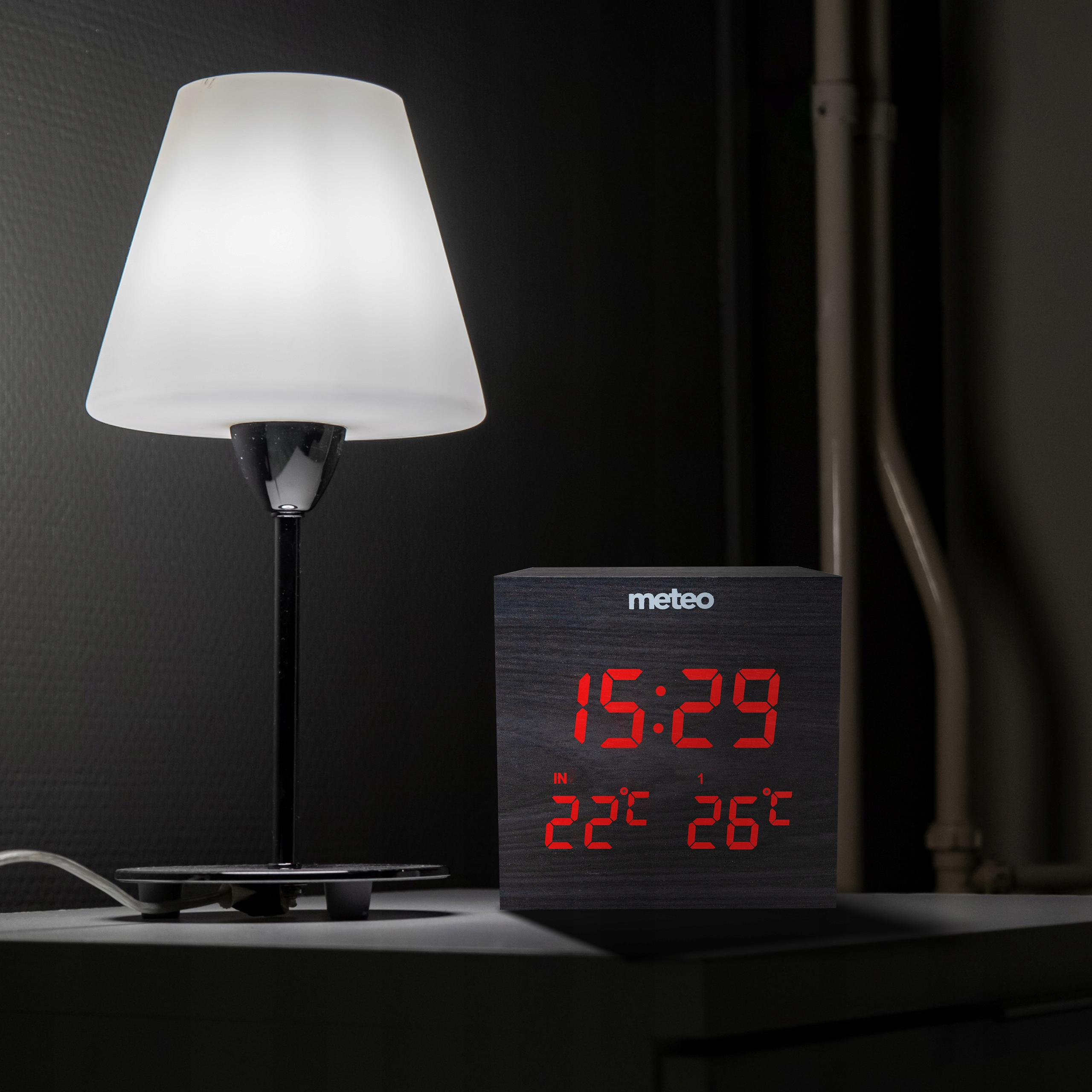 Zapalona lampa i cyfrowy zegar wyświetlający godzinę i temperaturę na ciemnej powierzchni. Czarna ściana w tle.
