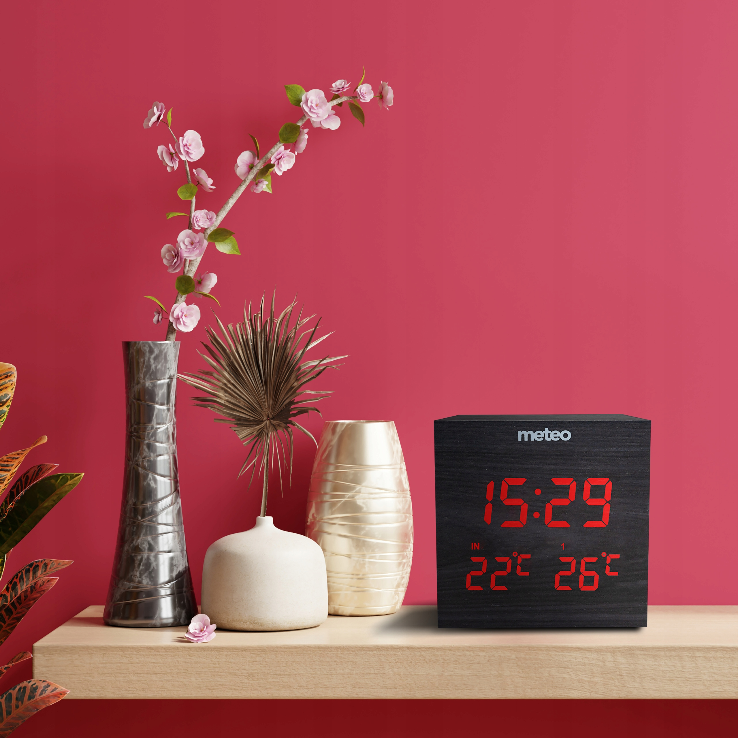 Zegar pokazuje 15:29 i temperaturę. Wazony i kwiaty na drewnianej półce na czerwonym tle.