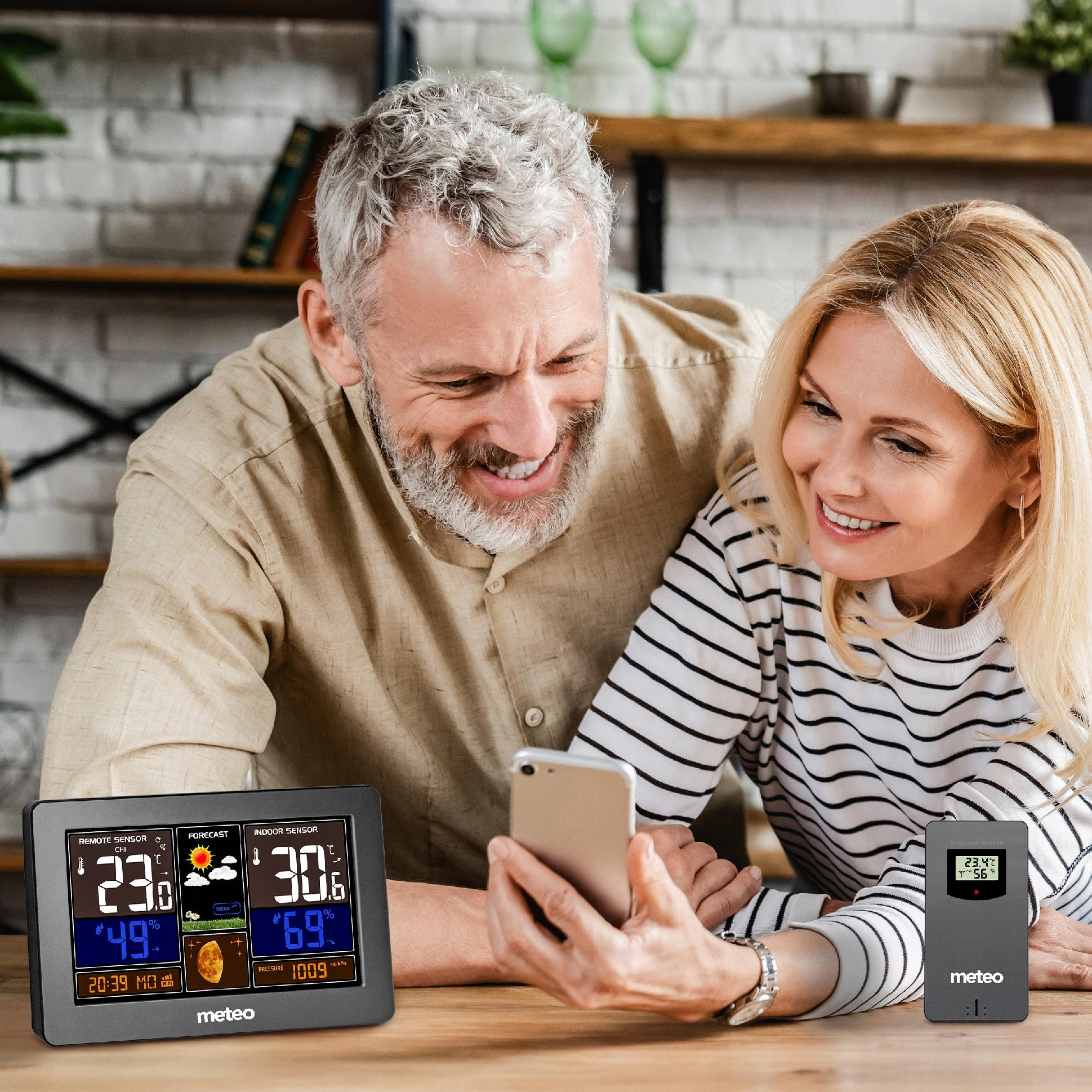 Para patrzy na telefon, uśmiechając się. Stacje pogodowe na stole. Mężczyzna ma beżową koszulę.