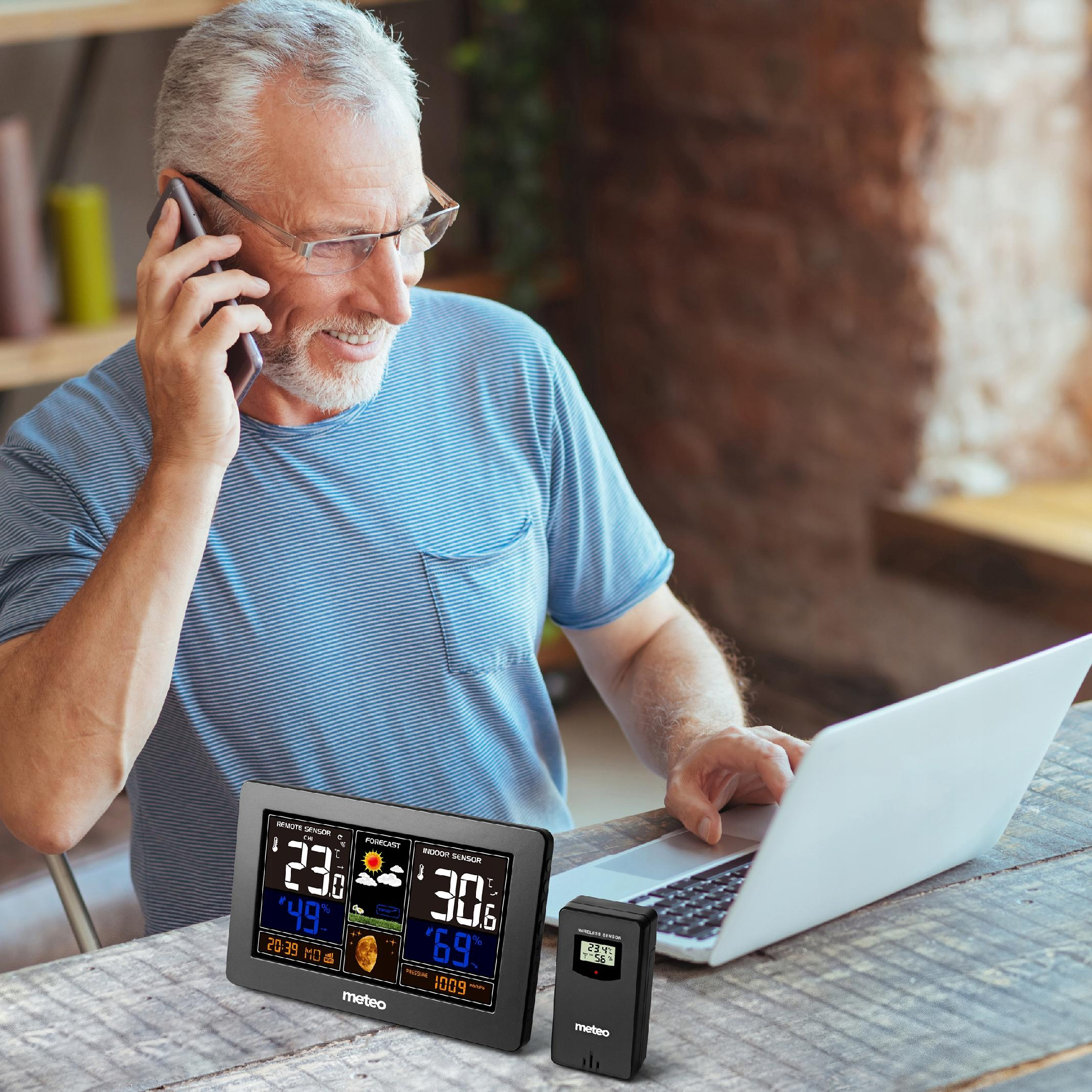 Mężczyzna w okularach rozmawia przez telefon, używa laptopa i ma stację pogodową na stole.