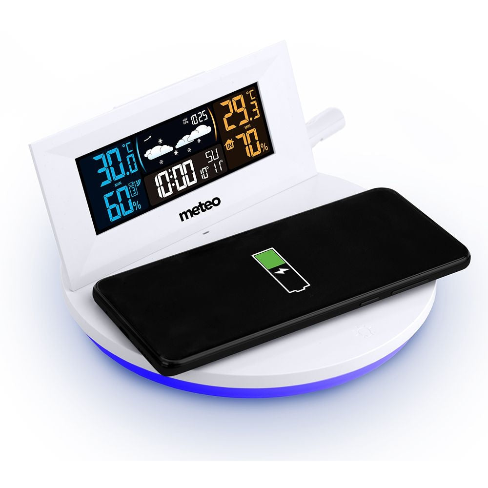 Biała cyfrowa stacja pogodowa i ładowarka do telefonu. Czarny telefon spoczywa na ładowarce. Stacja pokazuje temperaturę i czas.