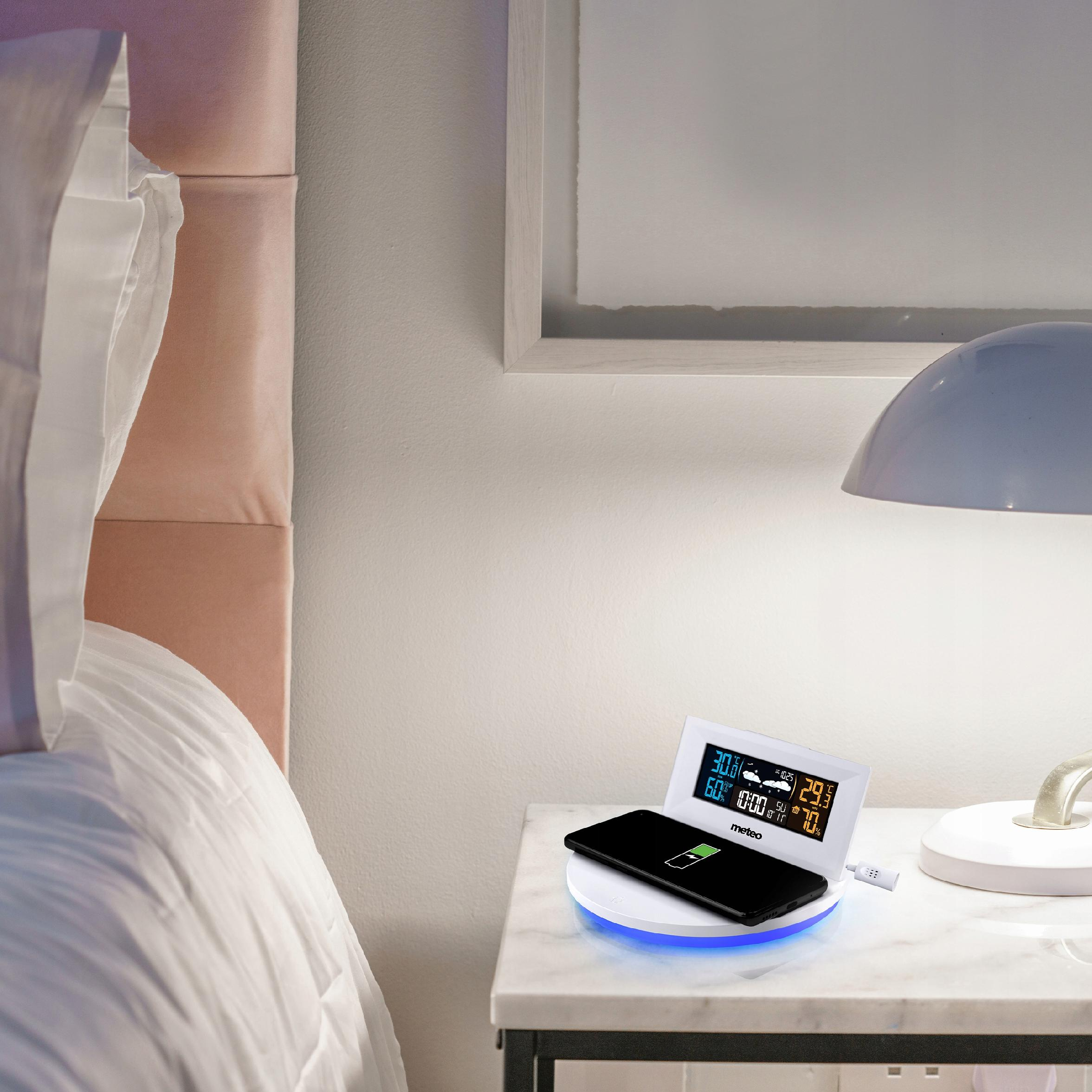 Szafka nocna z ładującym się telefonem, stacją pogodową i lampą. W tle łóżko.