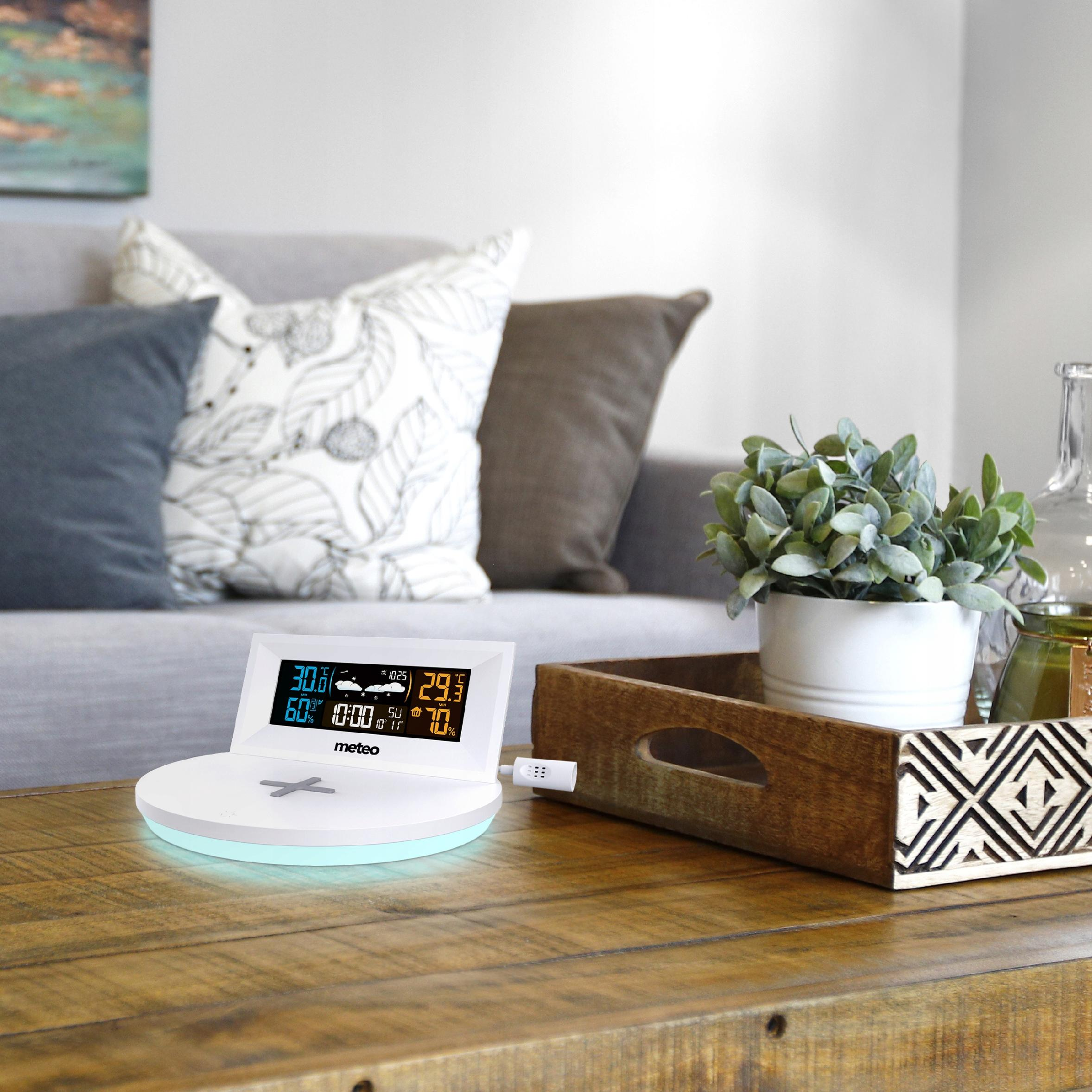 Stacja pogodowa wyświetla temperaturę i wilgotność na drewnianym stole, z rośliną i poduszkami w tle.