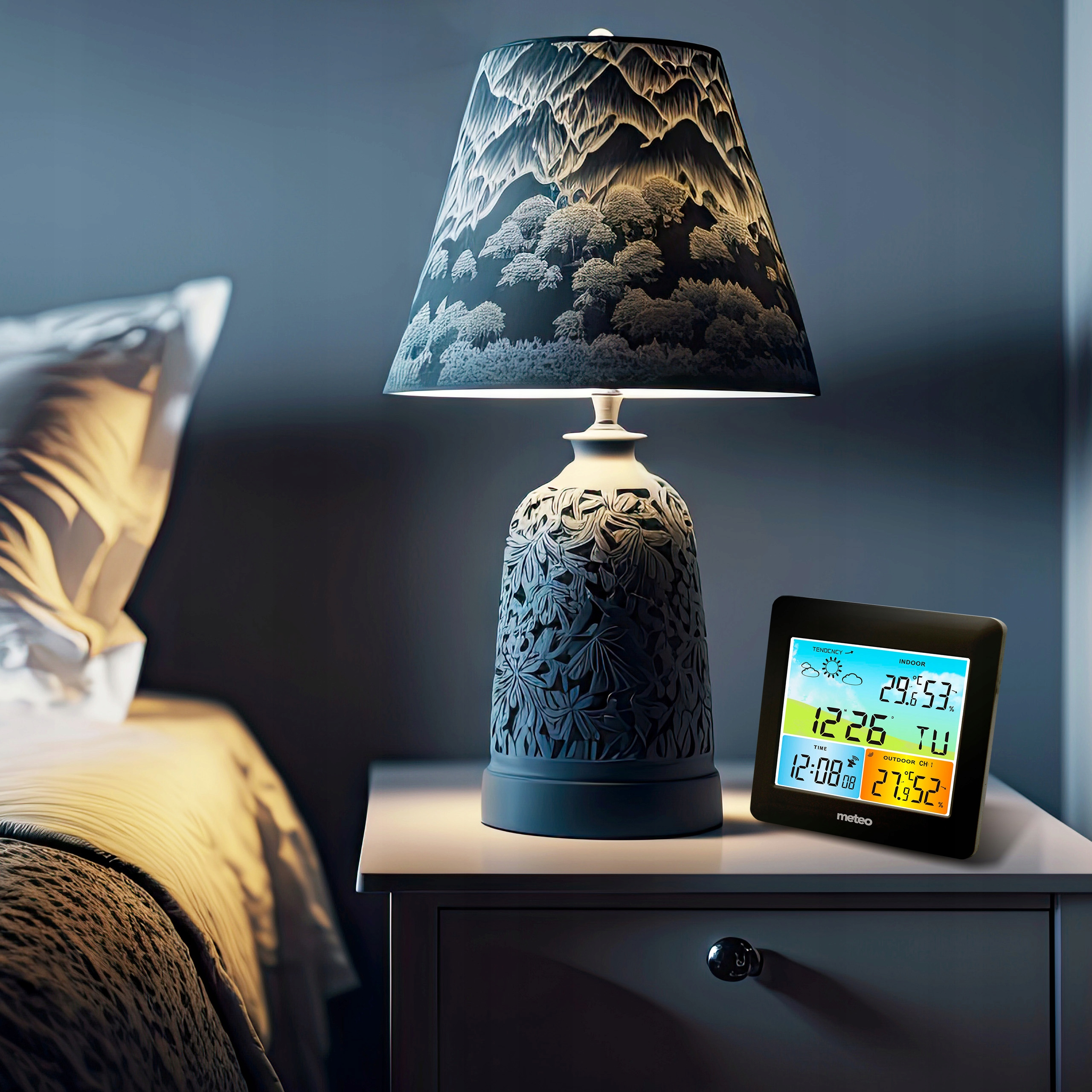 Szafka nocna z lampą i stacją pogodową. W tle łóżko.