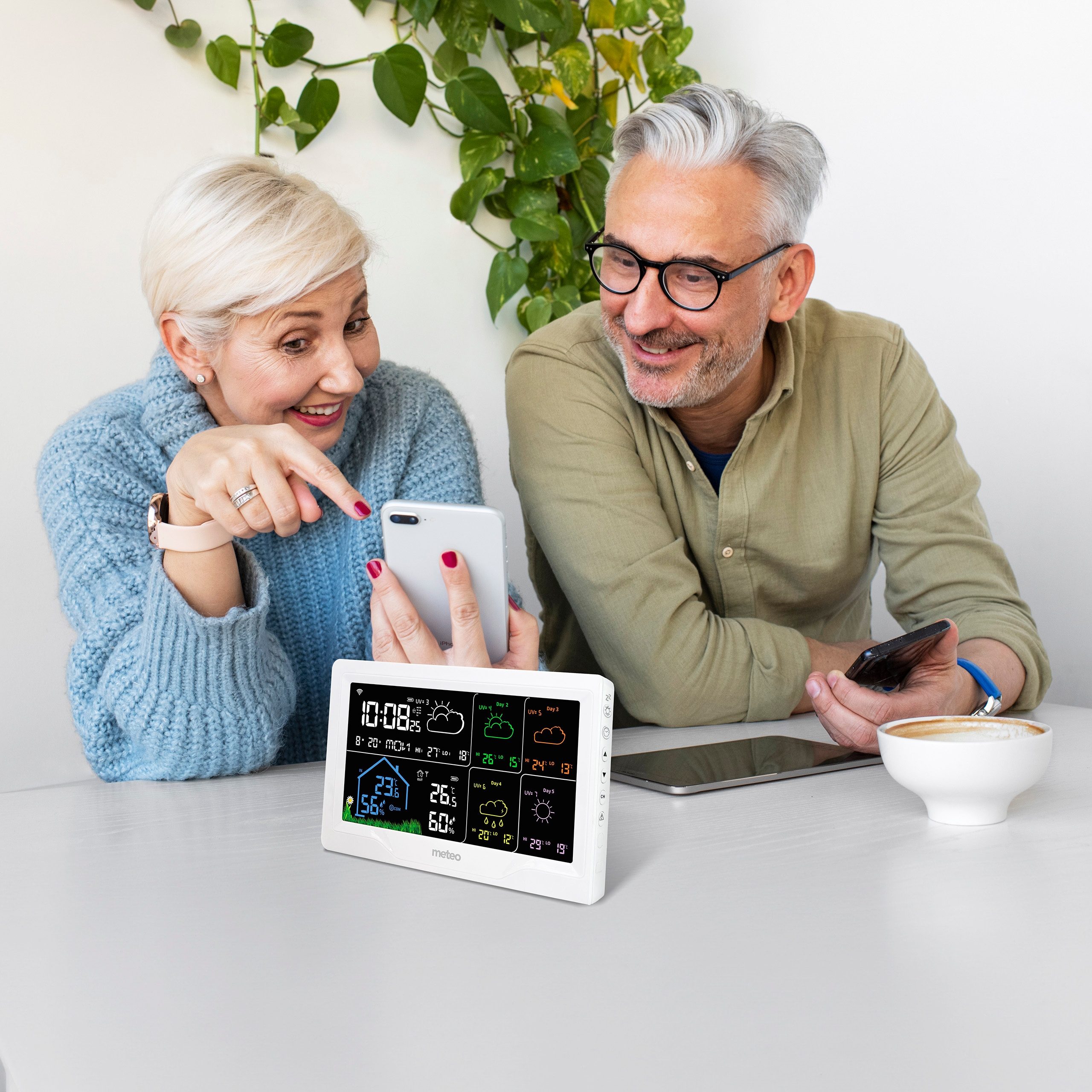 Starsza para patrzy na stację pogodową. Jedna osoba wskazuje na telefon, druga trzyma filiżankę kawy.
