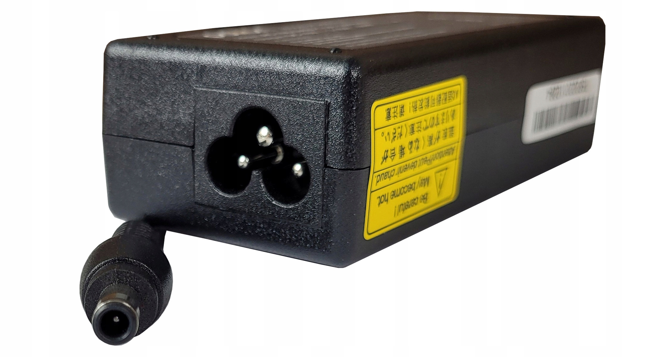 Czarny zasilacz do laptopa. Posiada wtyczkę trzybolcową, złącze beczkowe i żółtą etykietę ostrzegawczą. Tło białe.