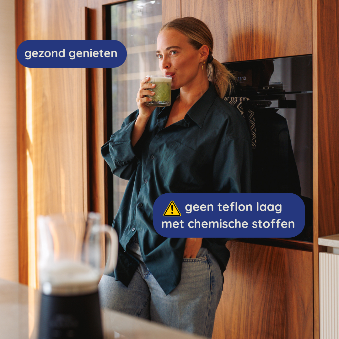Vrouw drinkt uit een groene drank in een glas. Ze draagt een donker overhemd en jeans, leunend tegen houten kasten.