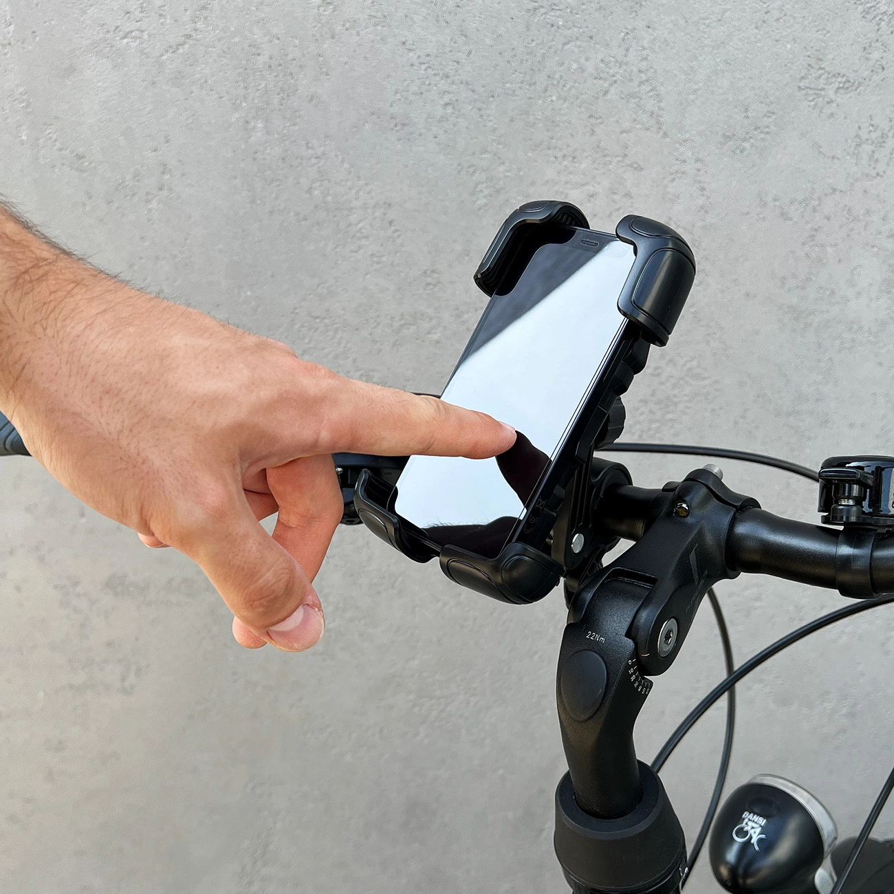 Eine Hand berührt ein Telefon in einer schwarzen Halterung an einem Fahrradlenker. Der Hintergrund ist eine graue Wand.