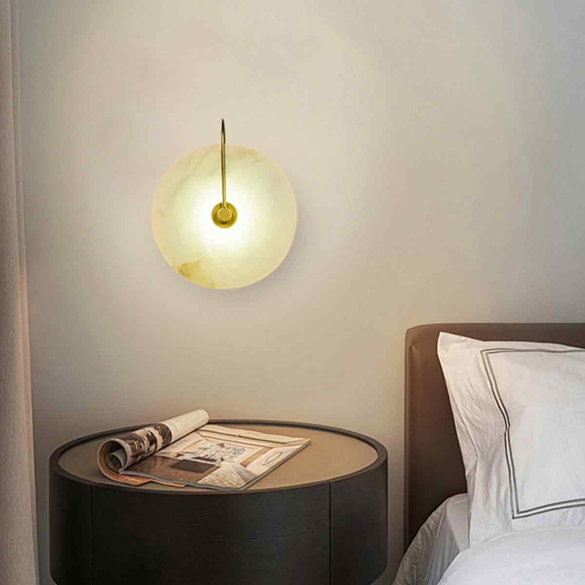 Eine runde Wandlampe beleuchtet eine Schlafzene. Ein Nachttisch hat eine Zeitschrift. Ein Bett mit weißen Kissen ist rechts.