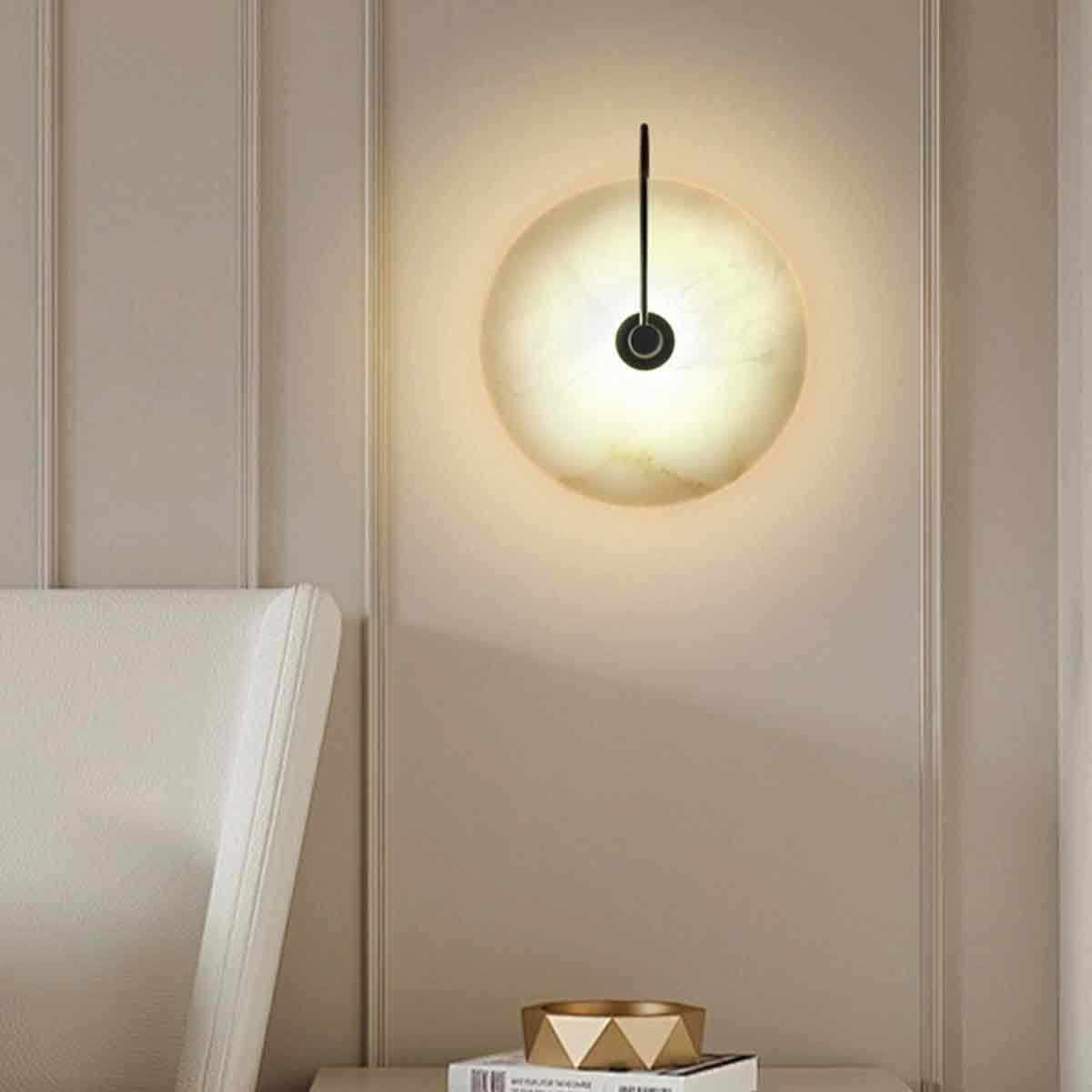 Eine runde Wandlampe strahlt warmes Licht aus. Eine schwarze Stange verbindet die Lampe mit der Wand. Links ist ein weißer Stuhl und darunter eine goldene Schale.