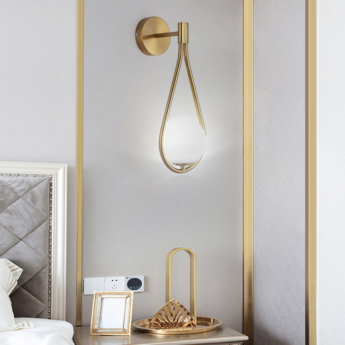 Eine goldene Wandlampe mit Tropfendesign. Ein Nachttisch hat ein Tablett, einen Bilderrahmen und Dekoration. Die Wand hat goldene Zierleisten.
