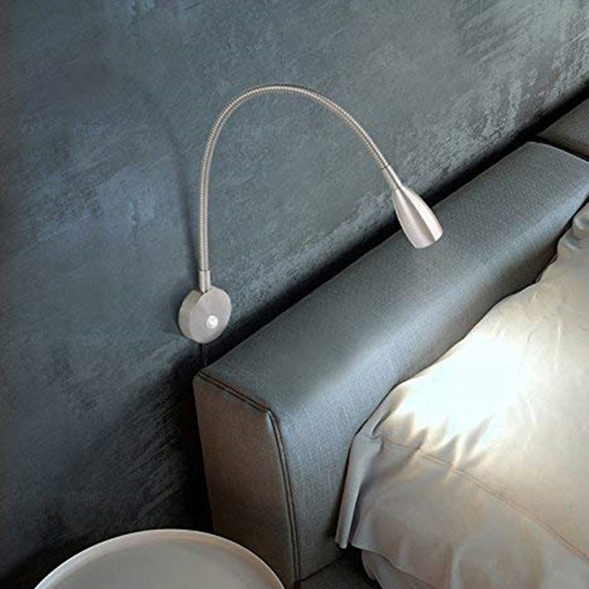Eine metallische Leselampe ist an einer strukturierten blauen Wand montiert. Rechts befindet sich ein Bett mit weißer Bettwäsche.