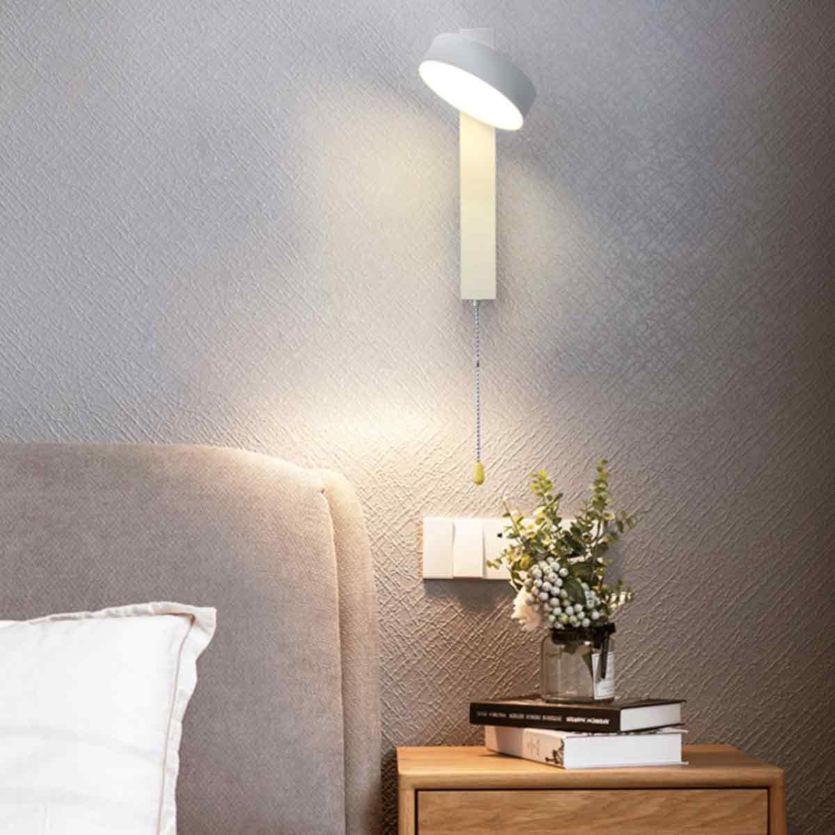 Ein modernes Schlafzimmer mit Wandlampe, Bett, Nachttisch, Blumen und Büchern. Die Lampe ist an. Die Wand ist grau.