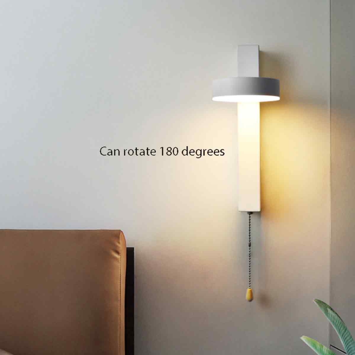 Eine moderne Wandlampe mit Kette und gelbem Zug. Sie ist an einer hellgrauen Wand montiert. Links ist ein Bettrahmen zu sehen.