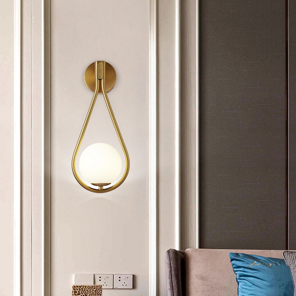 Eine goldene Wandleuchte mit Tropfendesign und rundem, weißem Licht. Sie ist an einer weißen Wand in der Nähe einer dunkelbraunen Wand angebracht.