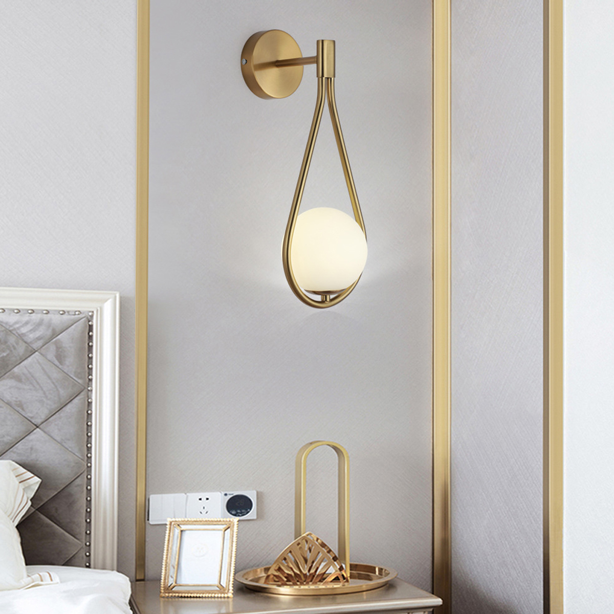 Eine goldene Wandlampe mit kugelförmigem Licht, Nachttisch mit Deko, Bilderrahmen und eine Wand mit goldenen Rahmen.