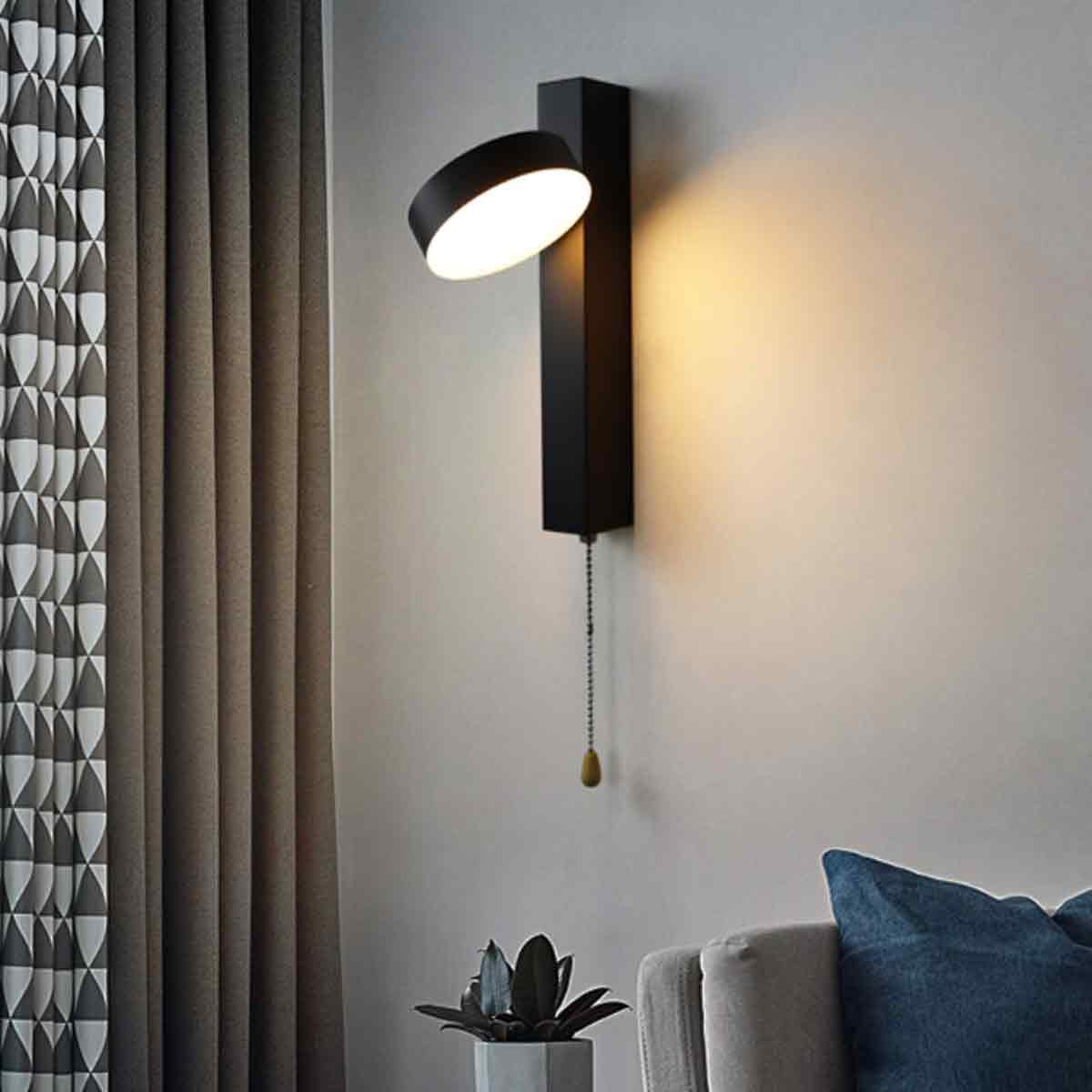 Eine moderne schwarze Wandleuchte mit Zugkette. Das Licht ist an und beleuchtet die Wand. Vorhänge und Sofa sind im Hintergrund zu sehen.