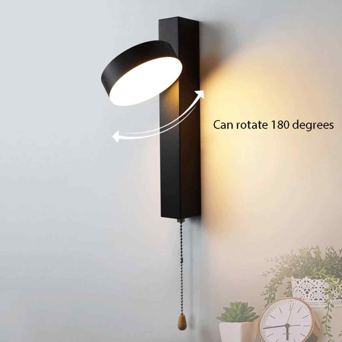 Schwarze Wandlampe mit rundem Licht. Sie kann sich um 180 Grad drehen. Eine Zugkette hängt unten. Uhr und Pflanzen unten.