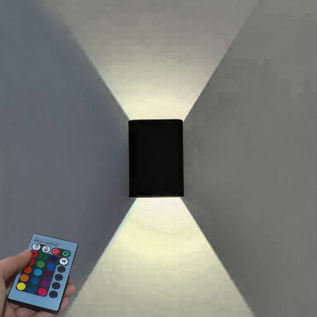 Eine schwarze Wandleuchte mit Lichtstrahlen. Eine Hand hält eine Fernbedienung mit bunten Knöpfen. Die Wand ist grau.