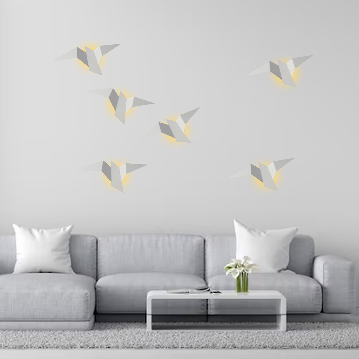 Wohnzimmer mit grauem Sofa, weißen Kissen und modernem Couchtisch. Dekorative vogelförmige Lichter an der weißen Wand.