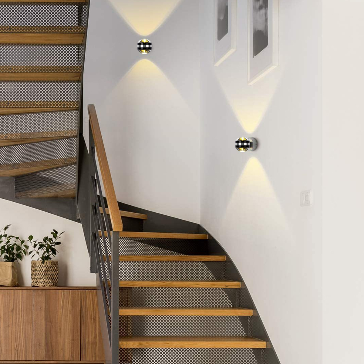 Holztreppe mit Metallgitter und Handläufen. Wandleuchten beleuchten die weiße Wand. Pflanzen und ein Holzschrank sind ebenfalls zu sehen.