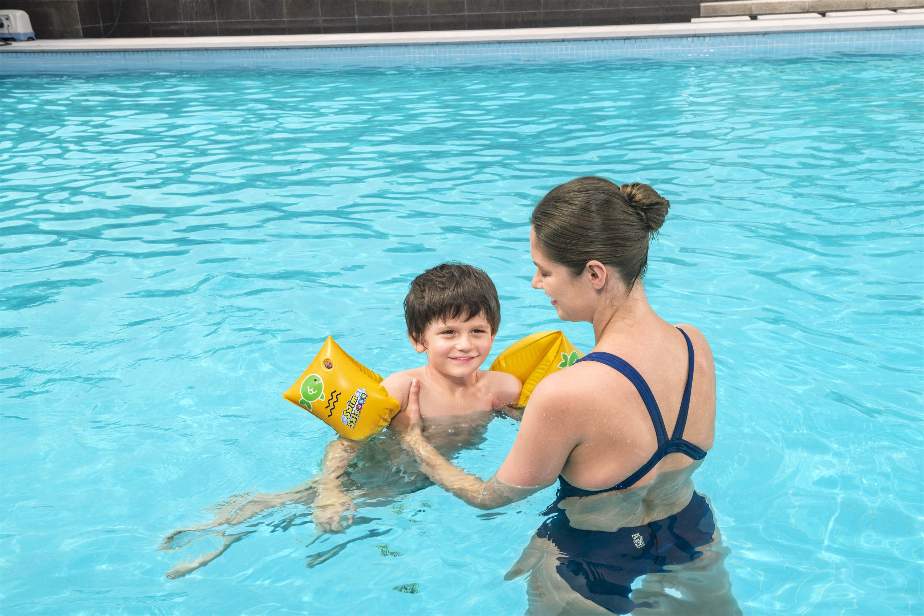 Eine Frau hilft einem Jungen im Pool. Der Junge trägt Armbänder. Beide sind im Wasser.