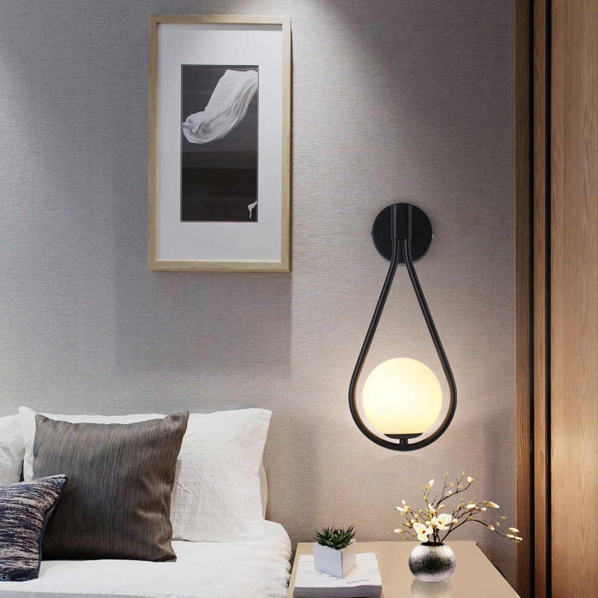 Ein Schlafzimmer mit Wandlampe, gerahmter Kunst und Bett. Die Lampe hat einen schwarzen Rahmen mit einem leuchtenden Globus. Pflanze und Blumen auf dem Nachttisch.