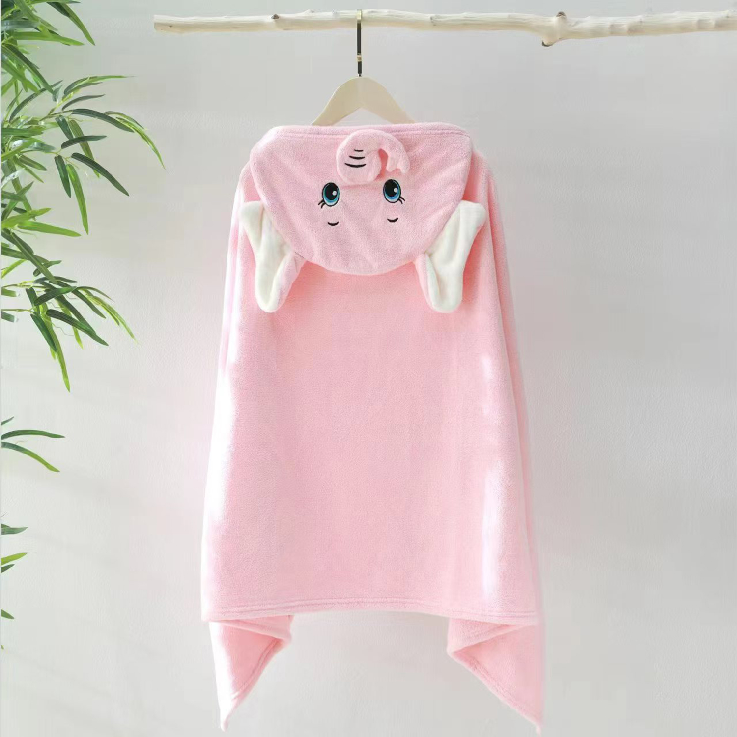 Un asciugamano rosa con cappuccio a forma di elefante è appeso a un appendiabiti di legno. Ha una faccia sorridente, orecchie e una proboscide. Foglie verdi sullo sfondo.