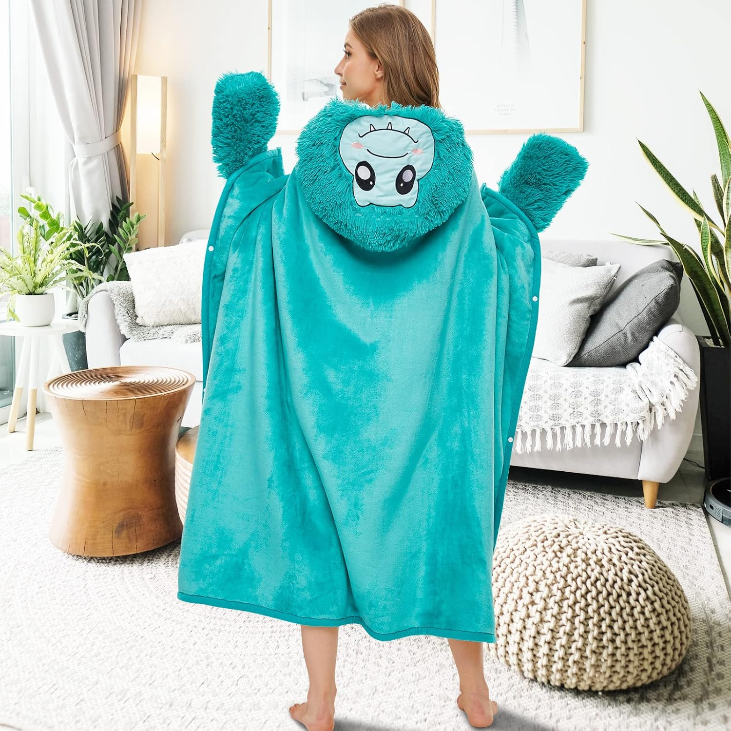 Una mujer con una manta con capucha turquesa con tema de monstruo. La escena es interior, con un sofá, mesa y alfombra visibles.