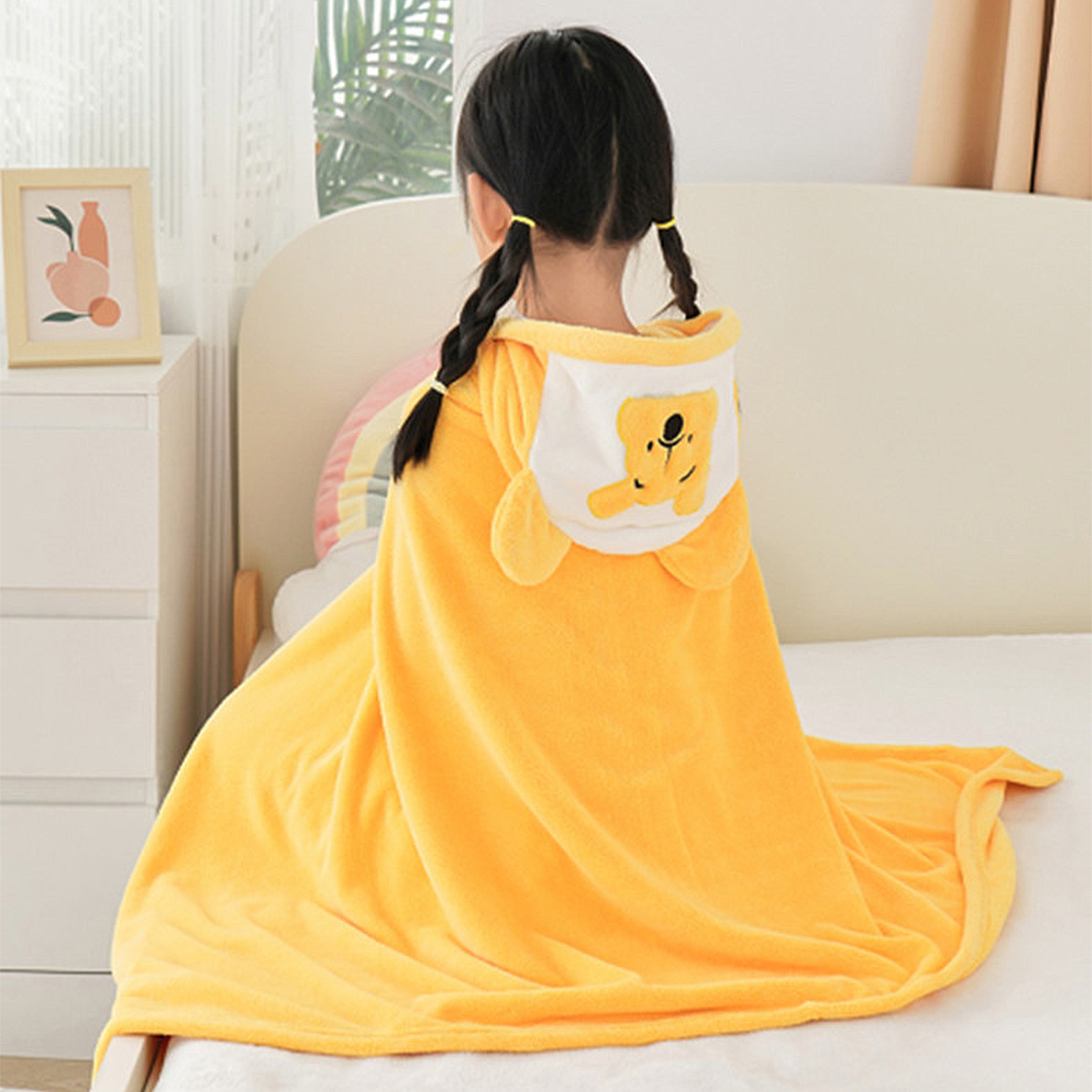 Una ragazza con trecce nere indossa un asciugamano giallo con cappuccio a forma di orso. È su un letto. Un quadro incorniciato e un armadietto bianco sono visibili.