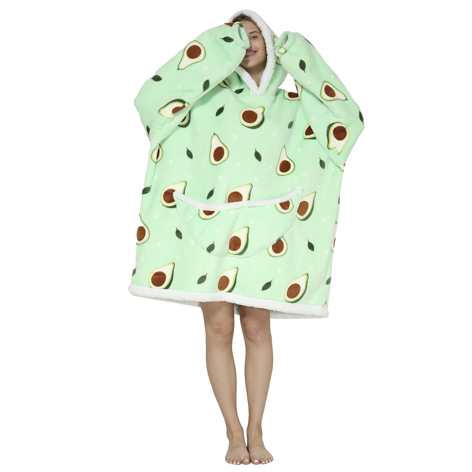 Una mujer de pie usa una sudadera con capucha estampada con aguacates. La sudadera es verde claro con aguacates y hojas. Sonríe con las manos en la cabeza.