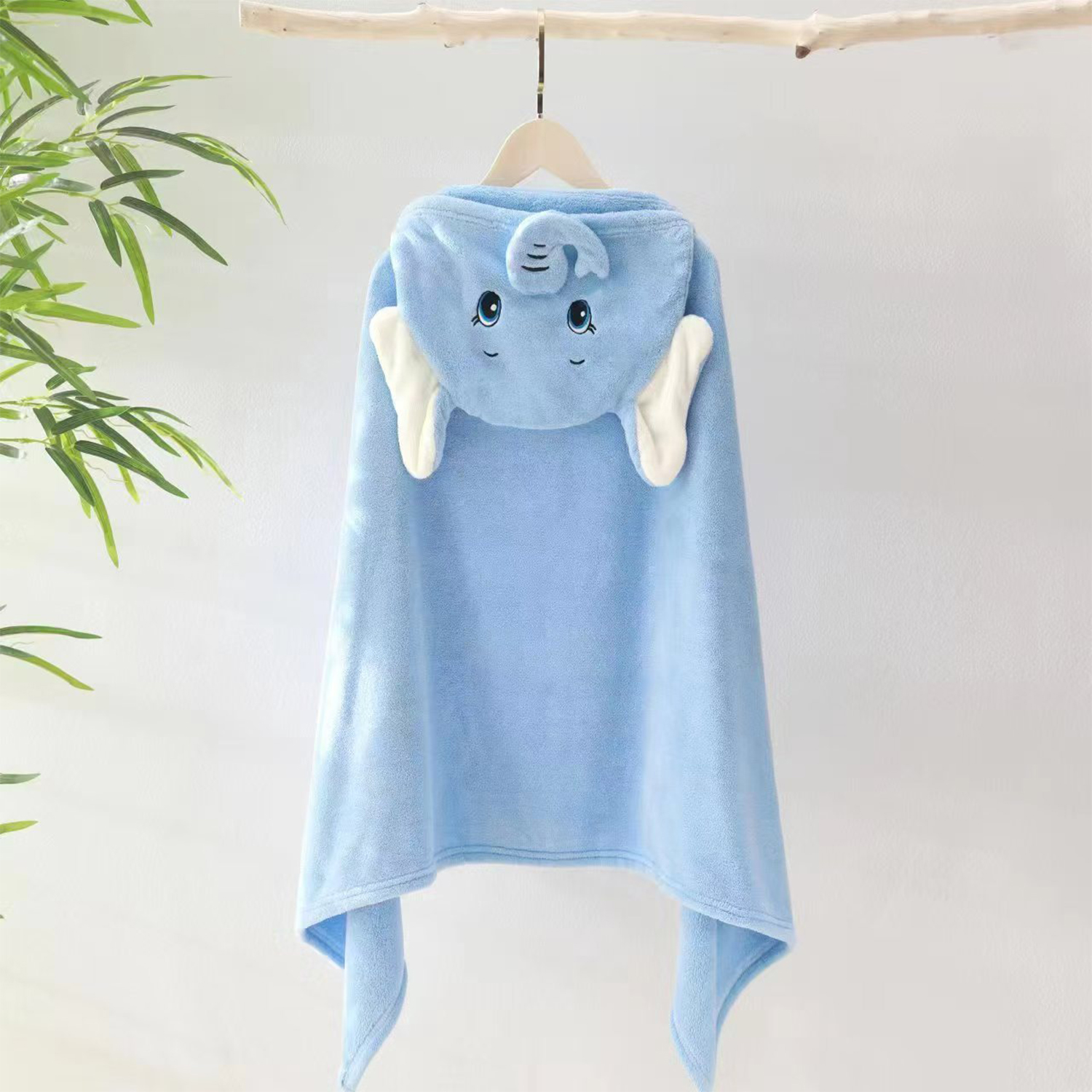 Una toalla con capucha azul con forma de elefante cuelga de una rama de madera. La toalla tiene una cara y orejas de elefante. Hay hojas verdes en el fondo.