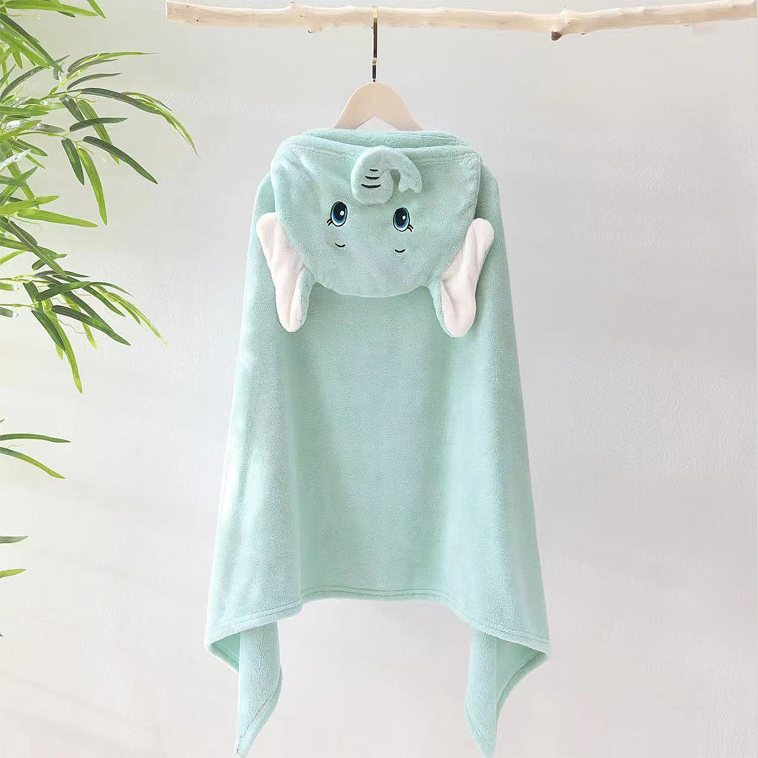 Una toalla con capucha de elefante verde menta cuelga de una rama de madera. La toalla tiene orejas blancas y una cara sonriente.