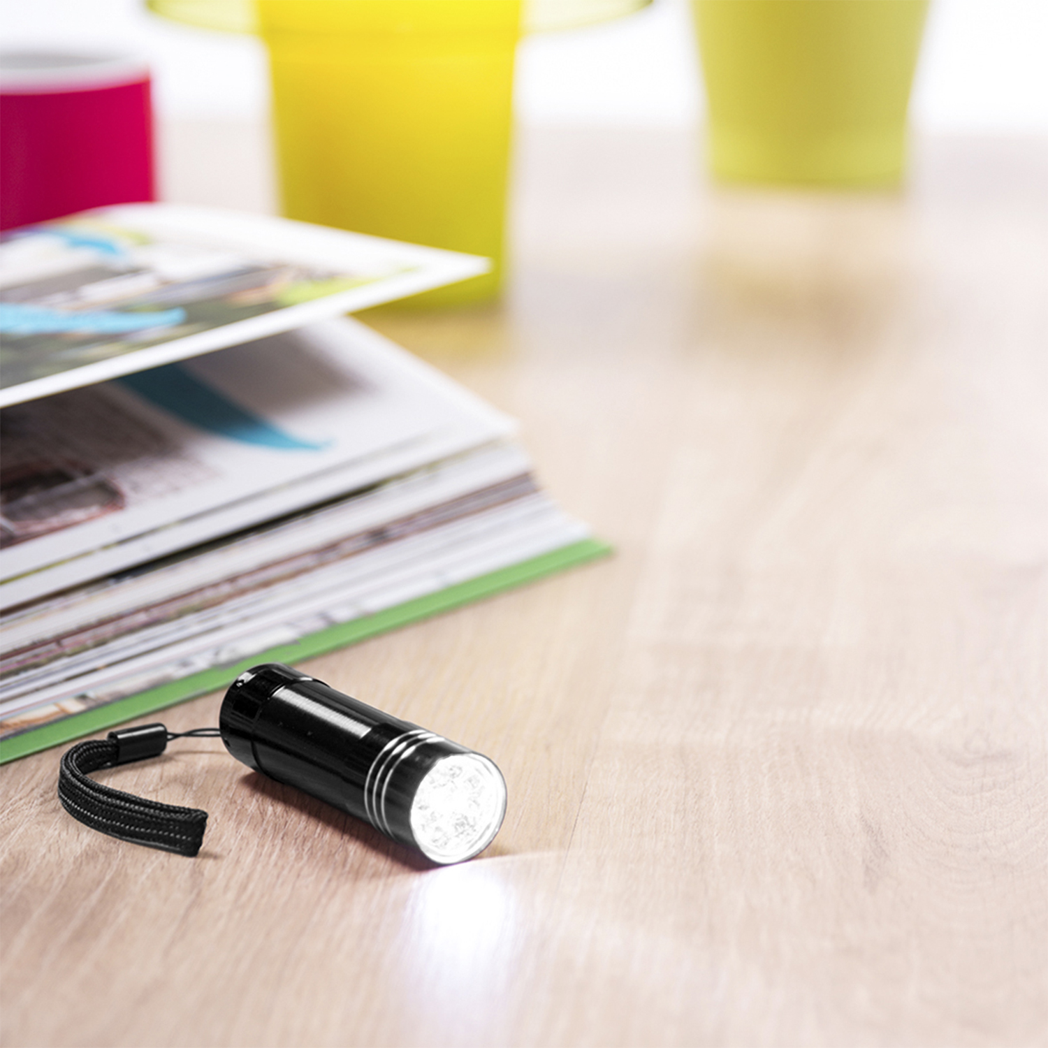 Una linterna negra sobre una superficie de madera, con revistas y tazas coloridas en el fondo. La linterna está encendida.