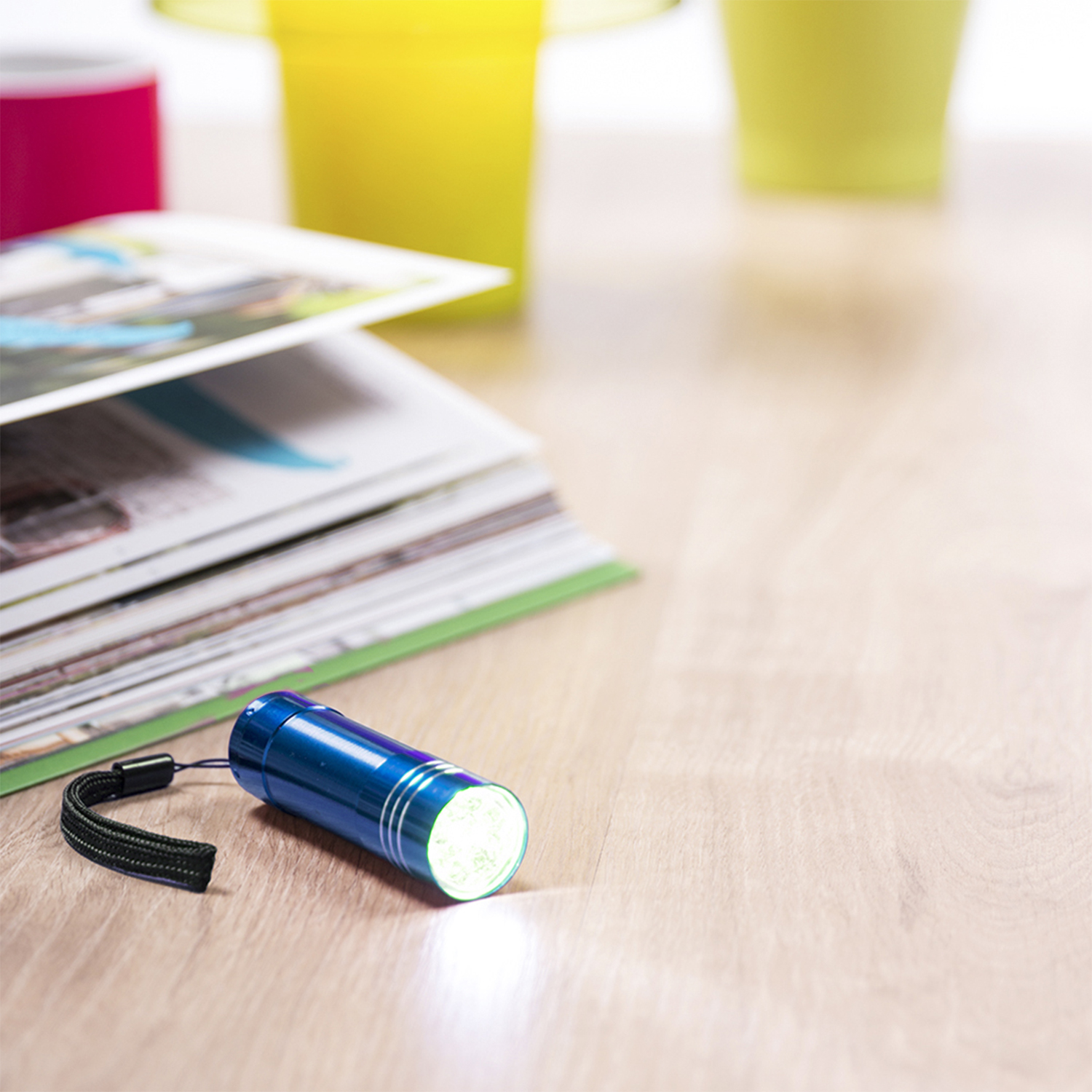 Una linterna azul con una correa negra está sobre una superficie de madera junto a revistas. Dos tazas de colores están en el fondo.
