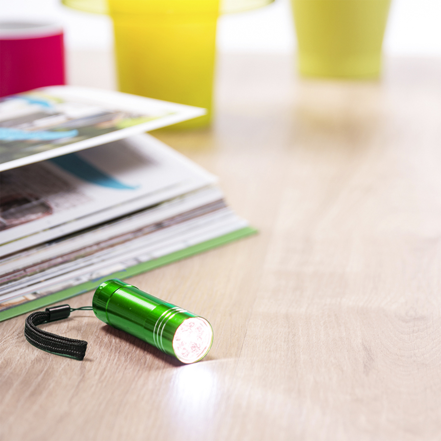 Una linterna verde está sobre una superficie de madera con una revista y objetos coloridos borrosos en el fondo.