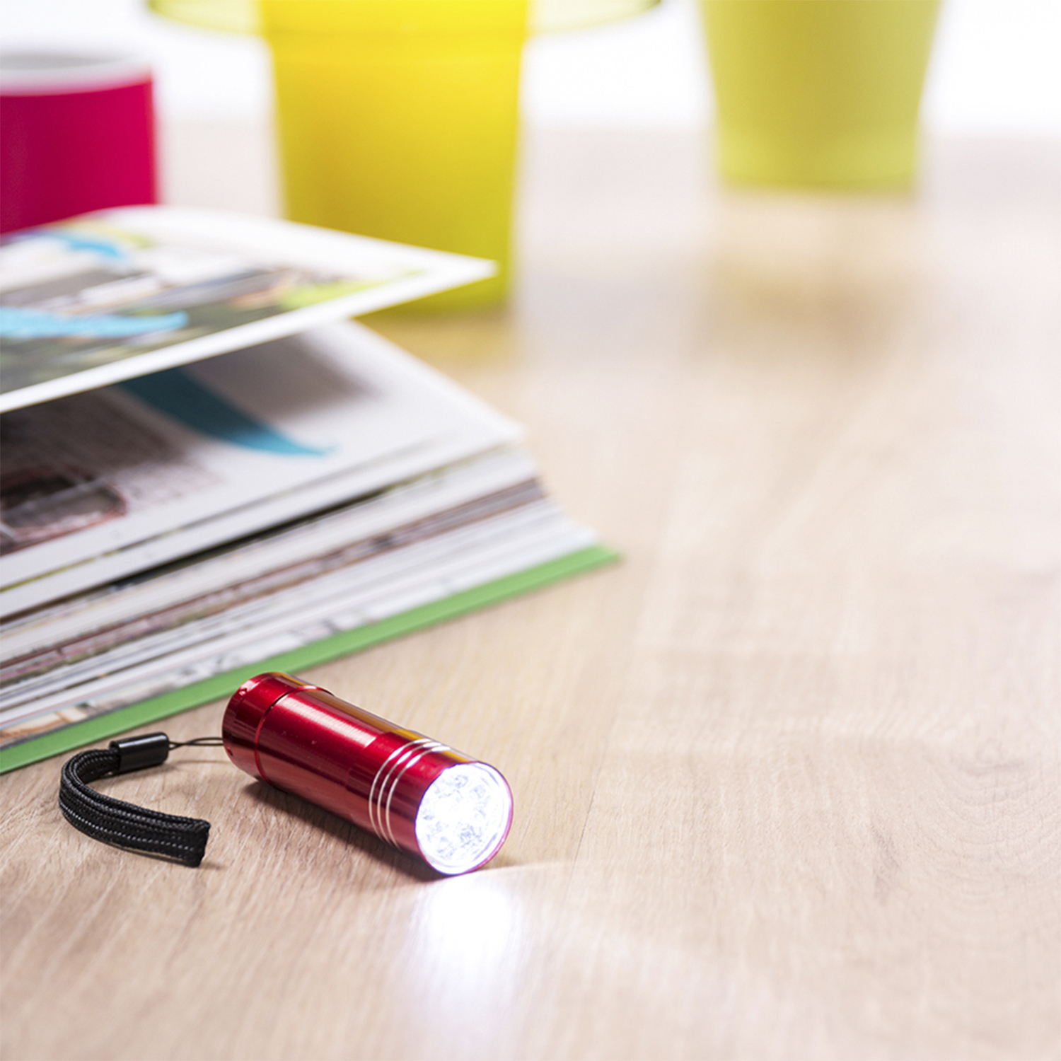 Una linterna roja con correa negra está sobre madera. Revistas y objetos coloridos al fondo.
