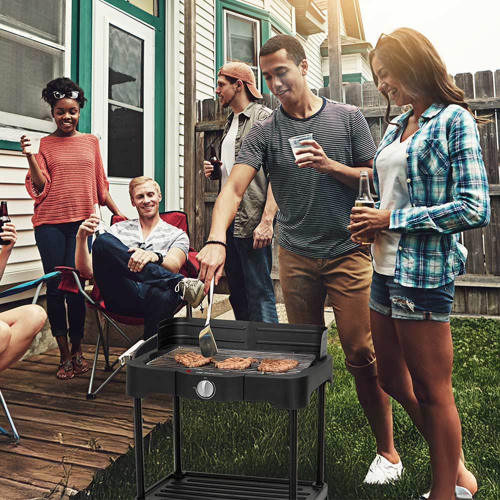 Grupo de amigos asando hamburguesas al aire libre. La gente está sosteniendo bebidas. Una parrilla negra está en primer plano, con hamburguesas cocinándose.