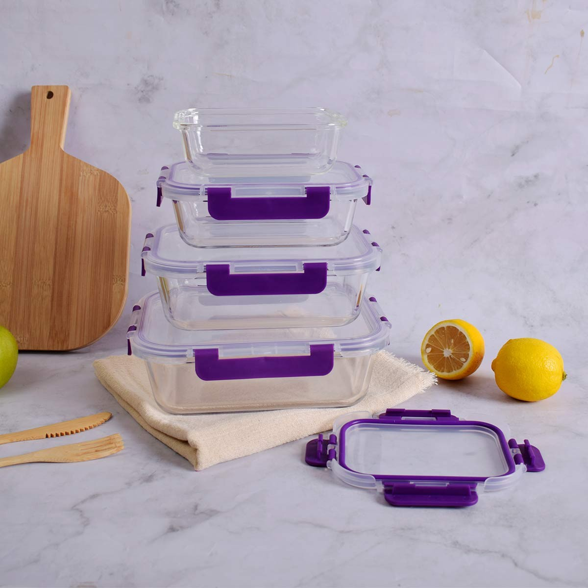 Pila de recipientes de vidrio para alimentos con cierres morados, limones, tabla de madera y utensilios sobre mármol.
