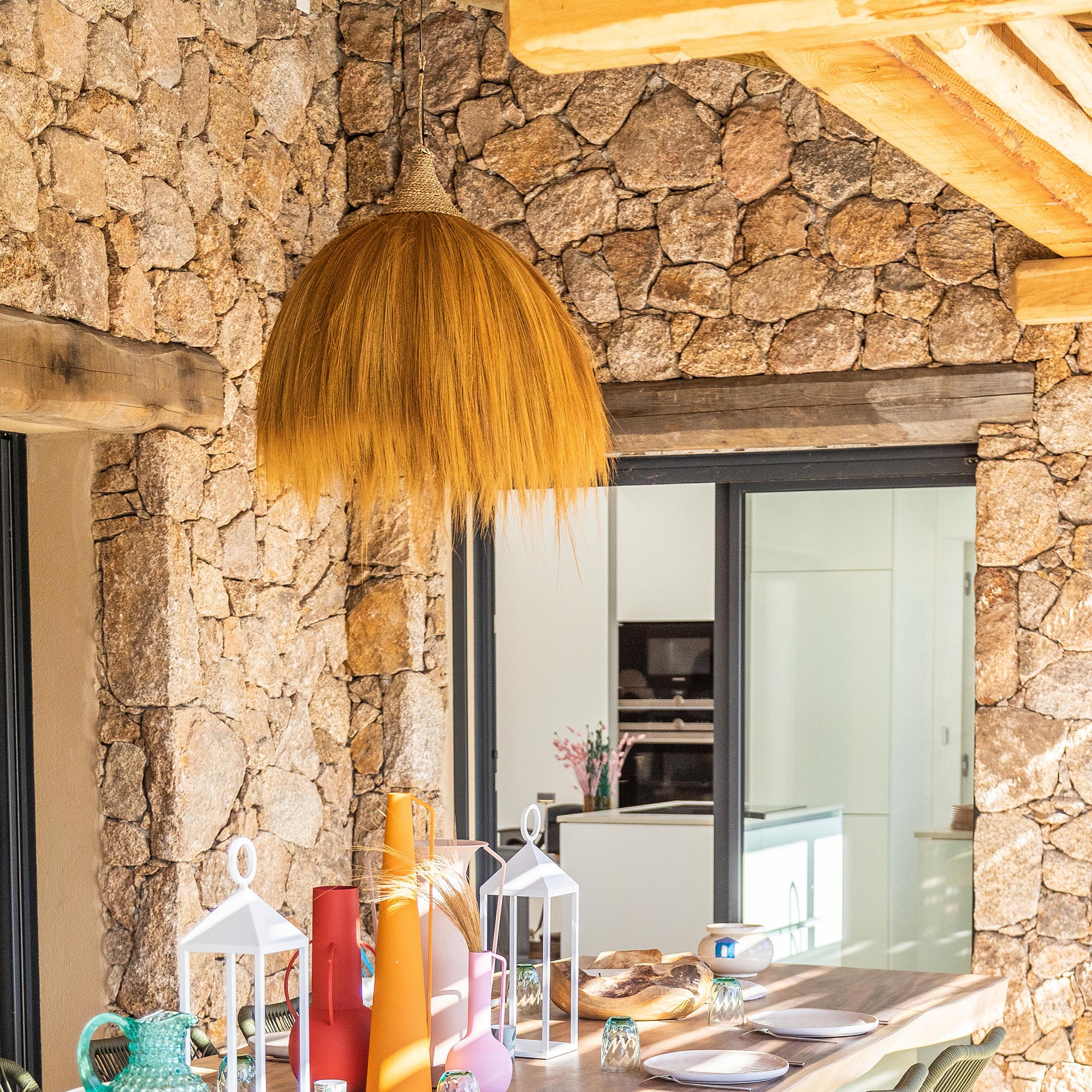 Szene im Freien. Eine Steinmauer hinter einem Holztisch mit bunten Vasen und Laternen. Überkopf hängt eine Strohlampe.