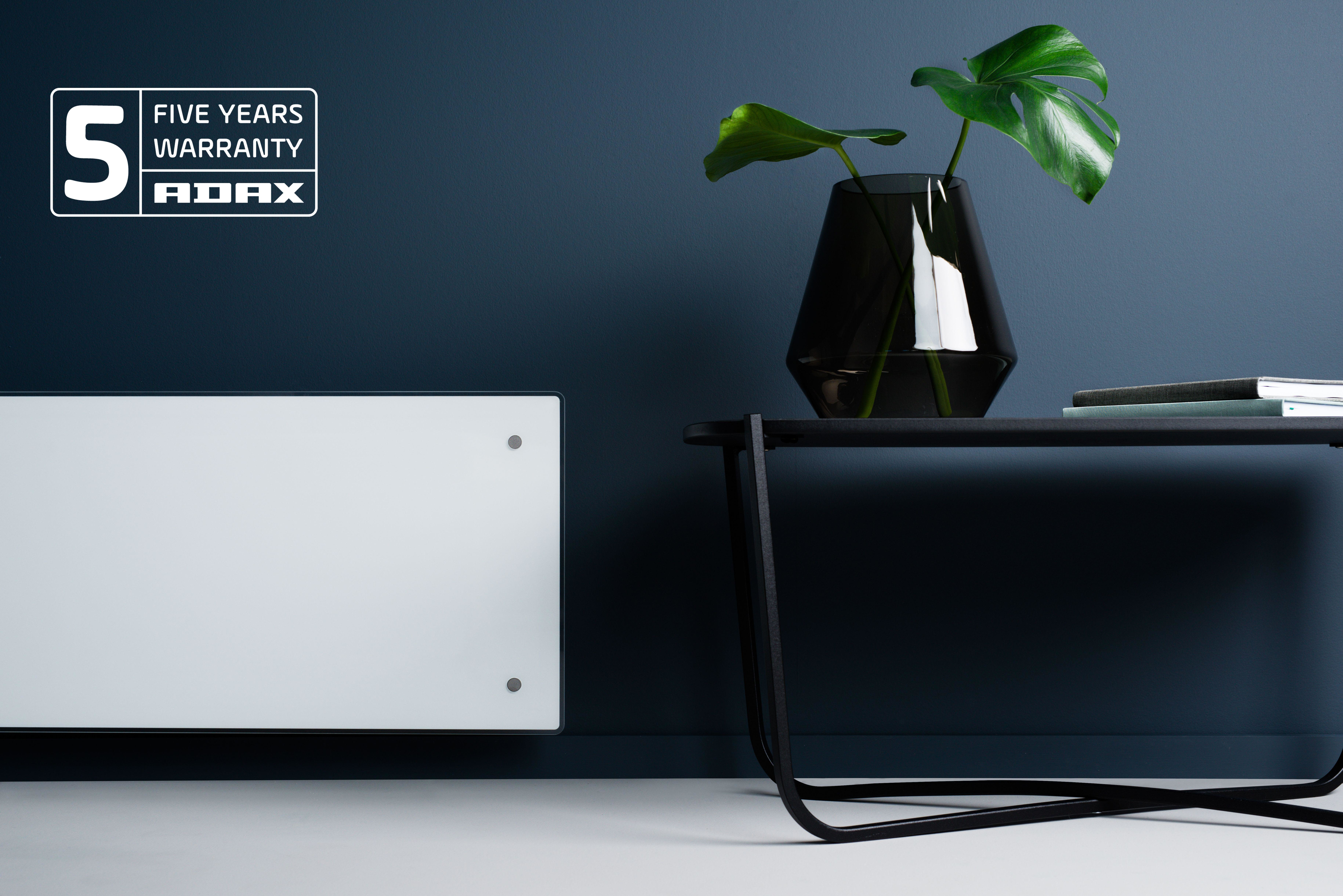 Un calefactor blanco en una pared blanca. Una mesa negra con jarrón, hojas y libros. Logotipo de garantía Adax.
