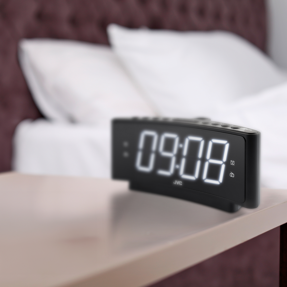 Czarny cyfrowy budzik JVC wyświetlający 09:08 stoi na stoliku nocnym. Rozmyte łóżko z białymi poduszkami i bordowym wezgłowiem.