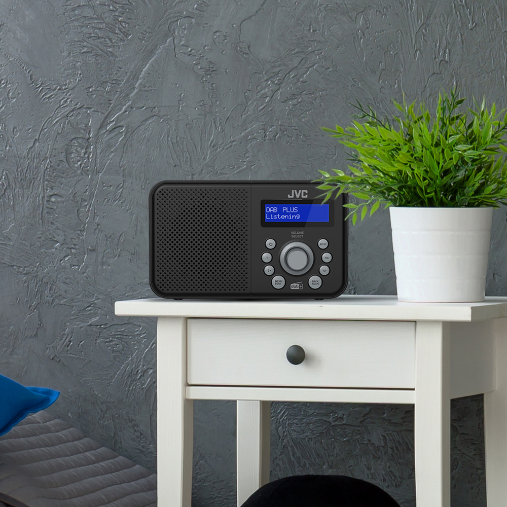 Czarny radio JVC stoi na białym stoliku. Zielona roślina w białej doniczce po prawej stronie. Tło to szara ściana.