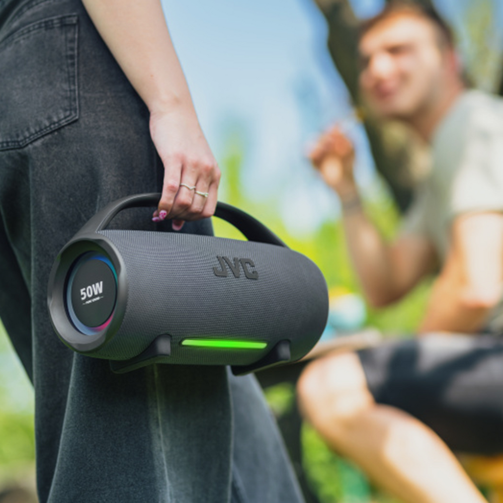 Osoba trzyma czarny głośnik JVC. Mężczyzna siedzi w rozmytym tle. Scena rozgrywa się na zewnątrz.