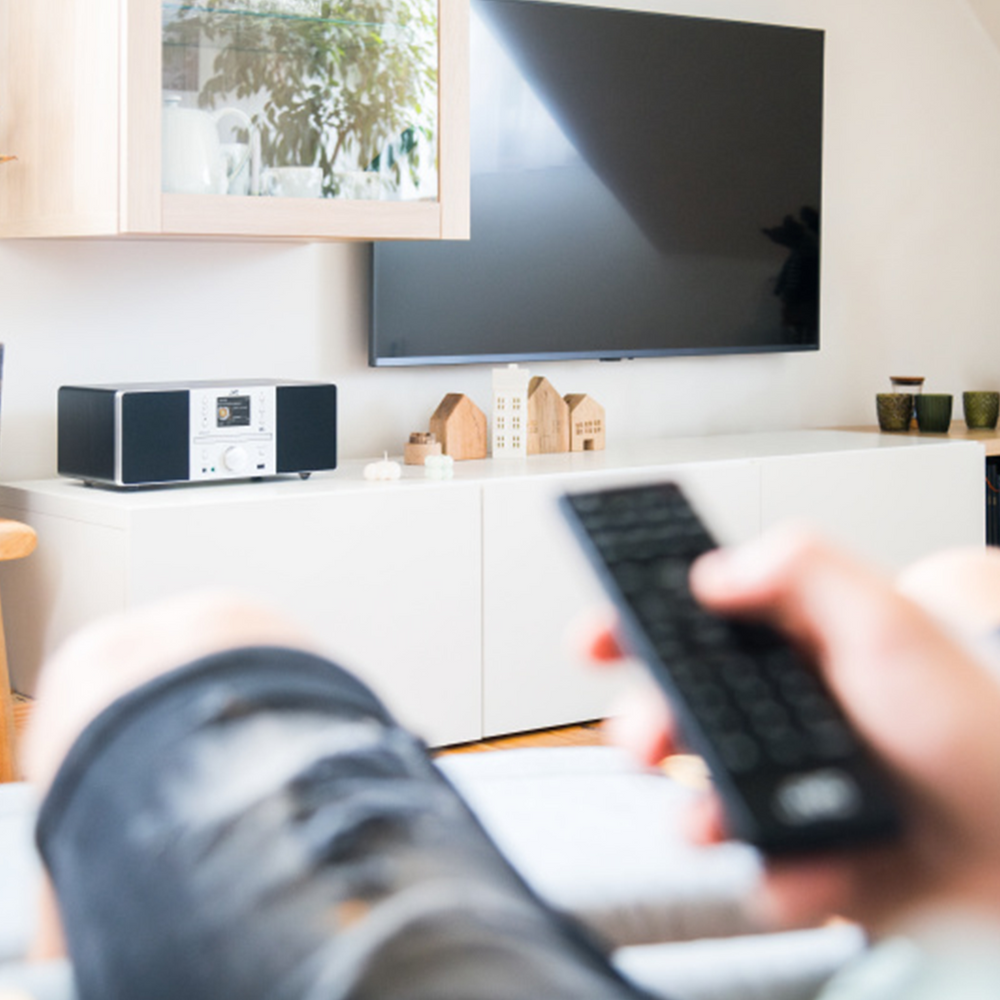 Osoba trzymająca pilota steruje telewizorem. Zestaw stereo i dekoracyjne domki stoją na białej szafce. Telewizor jest na ścianie.